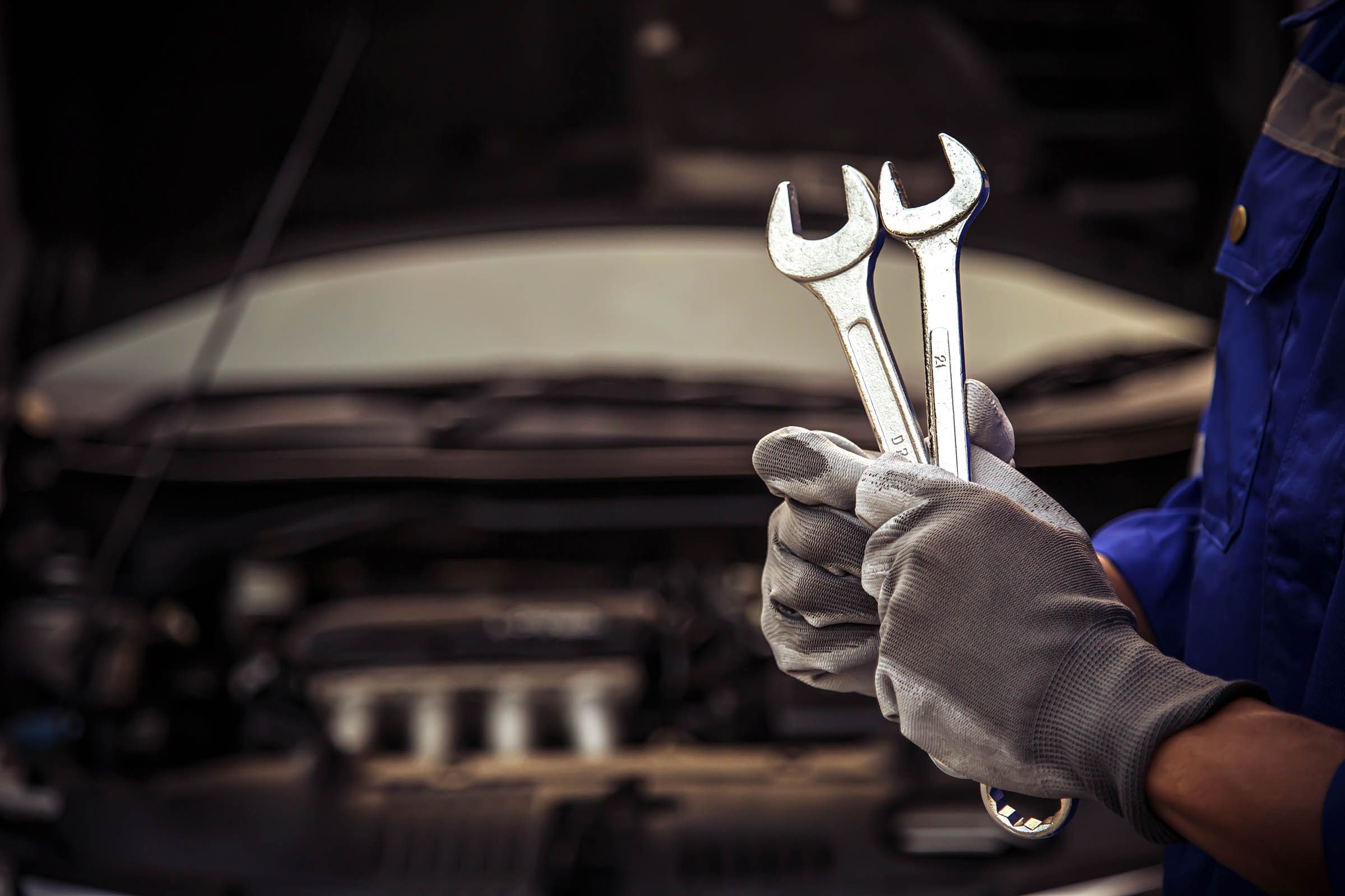 Eine Person in blauer Arbeitskleidung und grauen Handschuhen hält zwei verchromte Schraubenschlüssel in der Hand, während sie vor einem offenen Motorraum steht.